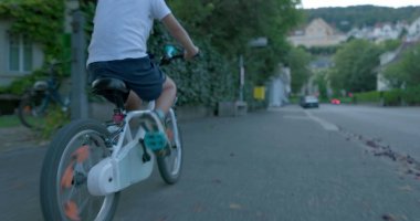 Beyaz bir bisiklete binen, şehir ortamında yaya geçidinden geçen, kasklı ve mavi çoraplı, güvenlik ve aktif yaşam tarzını vurgulayan genç bir çocuğun arka görüntüsü.