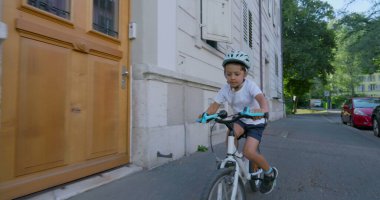 Beyaz kasklı ve beyaz gömlekli, şehir ortamında bisiklet süren genç bir çocuk, önündeki yola odaklanmış. Güneşli bir günün keyfini çıkarırken konut binalarının önünden geçiyor.