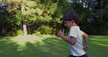 Parkta koşan genç bir çocuk, beyaz bir gömlek giymiş, kararlı bir ifadeyle, yeşilliklerle çevrili, güneşli bir günde dışarıda enerjik bir anın tadını çıkarıyor.