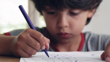 Genç çocuk yoğun bir şekilde renkli bir kalemle çizim yapmaya odaklanmış, ev ortamında bir masada otururken elindeki işe konsantre oluyor.