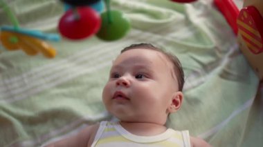 Bebek bir oyun hasırının üzerinde yatıyor, gözleri faltaşı gibi açık, renkli asılı oyuncakları inceliyor, oyuncu ve duyusal gelişimle meşgul, bir ev ortamında erken çocukluk keşfi.