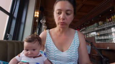 Anne ve bebek restoranda kucağında oturuyor, bebek çevreye bakıyor, anne yemeğe odaklanıyor, restoranda sıcak aile anı, bebek ve anneyle sıradan bir gezinti.