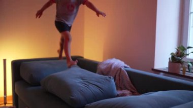 Genç bir çocuk kanepeye atlıyor, vücudunun düşmesine izin veriyor ve yastıkların üzerine düşüyor, akşam boyunca evde neşeli ve enerjik bir anın tadını çıkarıyor.