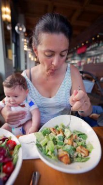Anne bir salata yerken ve bebeğini tutarken birden fazla iş yapıyor, bir restoranda günlük yemek sırasında bakıcılık ve yemek zamanını dengeliyor.