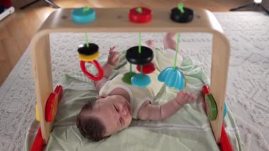 Bebek renkli oyuncakların altında oyun minderinde uzanıyor, duyusal nesnelere uzanıyor ve keşfediyor, erken gelişim aktiviteleri, sakin bir evde meraklı ve meşgul bir bebek.