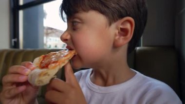 Küçük çocuk pizza diliminden büyük bir ısırık alıyor, ağzıyla peynir çekiyor, yan bakıyor ve aile dostu bir restoranda yemeğinin tadını çıkarmaya odaklanıyor.