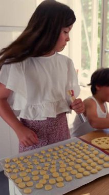 Genç bir kız hamur şekillerini pişirme tepsisine yerleştirirken abisi de yardım ediyor. İkisi de bir aile kurabiye yapma seansına katılıyor. Parçaları dikkatlice mutfağa yerleştirmeye odaklanıyor.