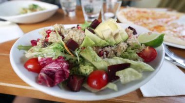 Yeşillikler, avokado, kiraz domatesleri, limon dilimleri ve bir restorandaki yemek masasında pancar kökü ile taze ve canlı bir kinoa salatası.