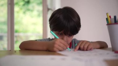 Genç bir çocuk elinde kalem tutarak bir masada resim çizmeye ve sanatına konsantre olmaya, sakin bir ev ortamında güzel motor beceriler ve yaratıcılık geliştirmeye odaklanmış.