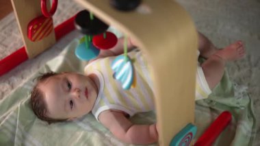 Bebek bir oyun hasırının üzerinde uzanıyor, renkli asılı oyuncakları gözlemliyor, şaşkın ve meraklı, sakin bir ev ortamında erken duyusal keşifler ve oyun zamanı.