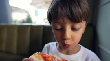 Küçük bir çocuk pizza diliminin tadını çıkarıyor, ağzına yakın tutuyor, gözleriyle yemeğe odaklanıyor, rahat bir restoranda rahat bir yemeğin tadını çıkarıyor.