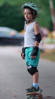 Genç bir çocuk kaykayın üzerinde güvenle duruyor, koruyucu giysi ve kask takıyor, eğlenceli ve güvenli bir oyun zamanında dışarıda sürmeye hazırlanıyor.