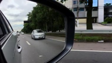 Otobanın yan aynasında, hareket halindeki araçları, ağaçları ve ilerideki yolu gösteren, çok şeritli bir yolda araba sürmenin deneyimini yakalayan bir görüntü.