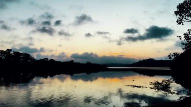 Gün batımında sakin bir göl, sakin bir su bulutları ve uzaktaki ağaçları yansıtıyor. Doğada huzurlu ve sakin bir manzara yaratıyor.