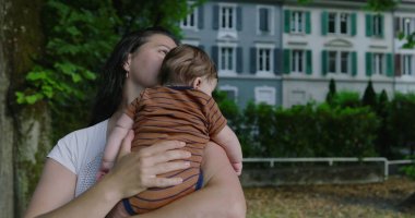 Anne, ağaçlarla çevrili bir şehir yolunda bebeğini tutarken huzur ve sevgi dolu bir ortamda konut binaları ile birlikte bir yere bakıyor.