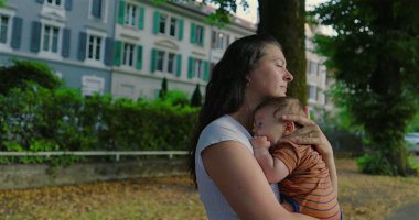 Anne, ağaçların çizdiği bir yolda bebeğini nazikçe kucaklıyor. Arkasında yeşillik ve binalar olan sakin bir şehir ortamında sıcaklık ve şefkat yayıyor.