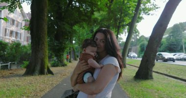 Anne, bebeği ağaçlık bir yolda tutuyor, kameraya bakıyor, arka plandan geçen arabalara bakıyor, kentsel bir ortamda huzurlu ve besleyici bir an sergiliyor.