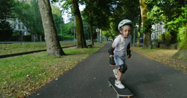 Çocuk kaykay yapıyor ağaçlarla kaplı bir yolda, miğfer ve koruyucu ekipmanlarla donatılmış, tahtada dengelenmiş, açık havada güneşli bir günün tadını çıkarıyor, yeşillik ve doğa ile çevrili.
