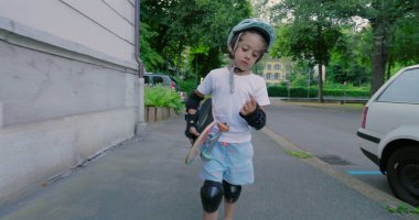 Şehir kaldırımında duran kaykaylı bir çocuk, kask ve koruyucu giysiler giyiyor, açık hava kaykay seansından sonra düşünceli görünüyor, çevresi yeşilliklerle çevrili.