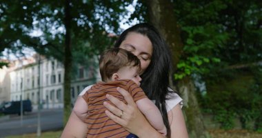 Anne, bebeğini ağaçlarla kaplı bir şehir yolunda tutarken sıcacık gülümsüyor yeşil ve yerleşim yerleriyle çevrili bir şefkat ve şefkat anı sergiliyor.