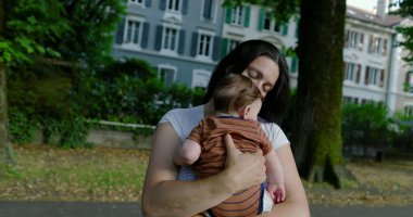 Anne, bebeğini ağaçlarla kaplı bir caddede kucaklıyor, derin sevgi ve özen gösteriyor, arka planda kentsel çevre ve yapraklı ağaçlar, huzurlu bir aile bağı anı.