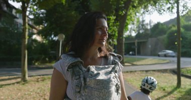 Ağaçlarla kaplı bir yolda bebeğiyle birlikte yürüyen, yan tarafa bakan, ağaçlarla çevrili huzurlu bir şehir parkı ve arka planda oynayan uzak bir çocuk.