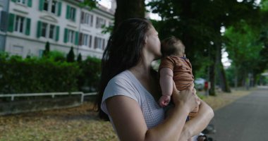 Anne, ağaçlarla çevrili bir şehir yolunda bebeğini tutarken huzur ve sevgi dolu bir ortamda konut binaları ile birlikte bir yere bakıyor.