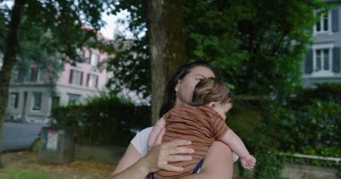 Anne, ağaçlarla çevrili bir şehir yolunda bebeğini tutarken huzur ve sevgi dolu bir ortamda konut binaları ile birlikte bir yere bakıyor.