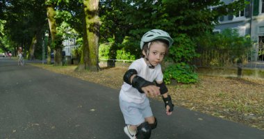 Çocuk kaykay yapıyor ağaçlarla kaplı bir yolda, miğfer ve koruyucu ekipmanlarla donatılmış, tahtada dengelenmiş, açık havada güneşli bir günün tadını çıkarıyor, yeşillik ve doğa ile çevrili.