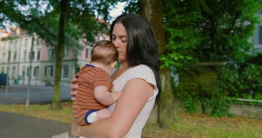 Anne, ağaçlarla çevrili bir yolda dikilirken bebeğini kucağına alıp sevgi ve şefkat gösteriyor. Arka planda şehir binaları ve ağaçlar var.