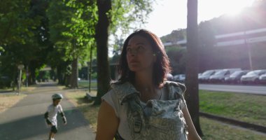 Ağaçlarla kaplı bir yolda bebeğiyle birlikte yürüyen, yan tarafa bakan, ağaçlarla çevrili huzurlu bir şehir parkı ve arka planda oynayan uzak bir çocuk.
