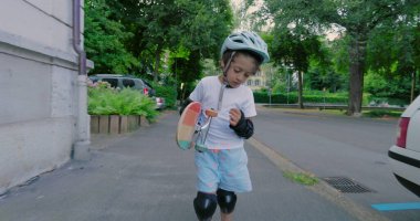 Şehir kaldırımında duran kaykaylı bir çocuk, kask ve koruyucu giysiler giyiyor, açık hava kaykay seansından sonra düşünceli görünüyor, çevresi yeşilliklerle çevrili.