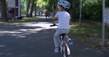 Bisikletli bir çocuk ağaçlık bir yolda sallanırken geri dönüyor, süper yavaş çekim, 800 fps, çocukluk eğlencesi, yaz boyunca kaskla parkta bisiklet sürüyor.