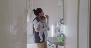 Anne bebeğini banyoda tutarken dişlerini fırçalıyor. Bebek, sabah rutinine ilgi göstererek çevreye merakla bakıyor.