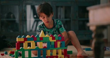 Genç çocuk bağdaş kurmuş, renkli ahşap blok yapısını gözlemliyor. Derin düşüncelere dalmış, elinde bir blokla bir sonraki hamlesini düşünüyor.