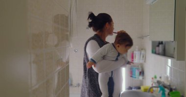 Anne, bebeğini banyoda taşırken dişlerini fırçalarken birden fazla iş yapıyor. Bebek, lavabodaki nesneleri merakla inceleyerek öne doğru eğilir.