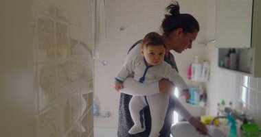 Anne, bebeğini banyoda taşırken dişlerini fırçalarken birden fazla iş yapıyor. Bebek, lavabodaki nesneleri merakla inceleyerek öne doğru eğilir.