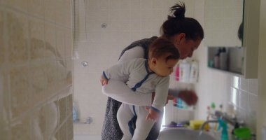 Anne, bebeğini banyoda taşırken dişlerini fırçalarken birden fazla iş yapıyor. Bebek, lavabodaki nesneleri merakla inceleyerek öne doğru eğilir.