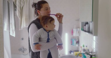 Anne bebeğini banyoda tutarken dişlerini fırçalıyor. Bebek, sabah rutinine ilgi göstererek çevreye merakla bakıyor.