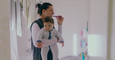 Anne banyoda birden fazla iş yapıyor, bebeğini tutarken dişlerini fırçalıyor. Bebek, sabah rutininden büyülenmiş bir şekilde çevreye merakla bakıyor.