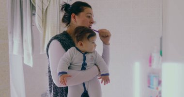 Anne banyoda birden fazla iş yapıyor, bebeğini tutarken dişlerini fırçalıyor. Bebek, sabah rutininden büyülenmiş bir şekilde çevreye merakla bakıyor.