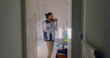 Anne banyoda birden fazla iş yapıyor, bebeğini tutarken dişlerini fırçalıyor. Bebek, sabah rutininden büyülenmiş bir şekilde çevreye merakla bakıyor.