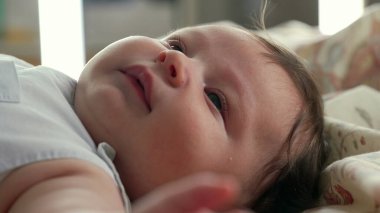 Bebek yatakta uzanıyor, belli belirsiz bir gülümsemeyle yukarı bakıyor. Yakından bakıldığında bebeğin yanakları ve yumuşak saçları vurgulanıyor. Bebek ise rahat ve rahat bir ortamda.