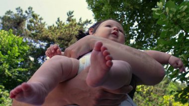 Bebeğini kucağında tutan bir annenin düşük açılı görüntüsü bebeğin kollarında rahatça dinlenirken ayaklarını ve bacaklarını gösteriyor, ağaçlarla çevrili.