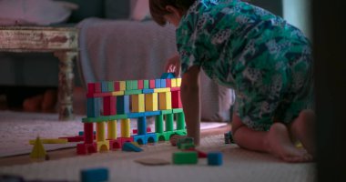 Genç bir çocuk yerde diz çökmüş, rahat bir oturma odası ortamında ahşap bloklarla renkli bir yapı inşa etmeye odaklanmış. Her bloğu dikkatlice yerleştirirken konsantrasyon ve yaratıcılık