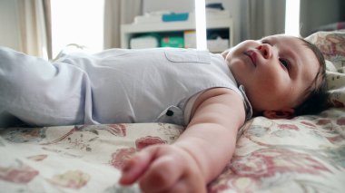 Bebek rahat bir şekilde battaniyeye uzanmış, tek kolla uzanıyor. Sahne, rahat bir evde dinlenirken bebeğin tüm vücudunu yakalıyor.