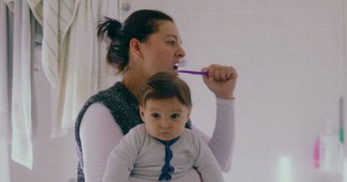 Anne banyoda birden fazla iş yapıyor, bebeğini tutarken dişlerini fırçalıyor. Bebek, sabah rutininden büyülenmiş bir şekilde çevreye merakla bakıyor.