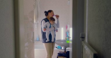 Anne banyoda birden fazla iş yapıyor, bebeğini tutarken dişlerini fırçalıyor. Bebek, sabah rutininden büyülenmiş bir şekilde çevreye merakla bakıyor.
