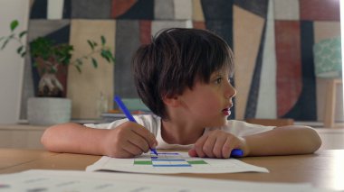 Genç bir çocuk, mavi bir kalemle bir meydanı boyuyor, bir masada yaratıcı aktivitelere odaklanıyor, konsantrasyon ve sanatsal gelişim gösteriyor.