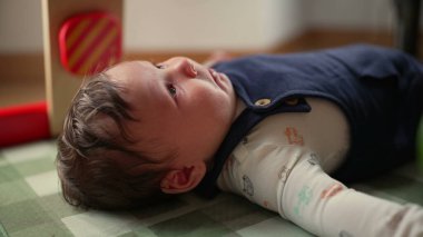 Bebek rahat bir evde paspasın üzerinde uzanıyor. Rahatsızlık ve yansıma gibi ince duygular gösteriyor. Bebek sakin görünüyor ama doğal ışıkta ilgiye ihtiyaç duyduğunun erken belirtilerini gösteriyor.